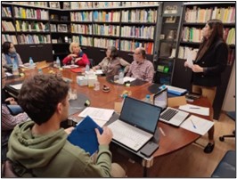 Varias personas se encuentran reunidas en torno a una gran mesa de reuniones. Sobre ella hay carpetas y ordenadores. Tras ellos se alza una gran estantería con libros. 